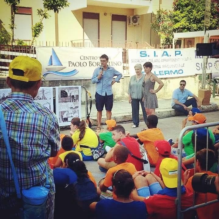 Luca parla al microfono a un gruppo di ragazzi con cappellini colorati, davanti al Centro Paolo Borsellino di Palermo, con le tavole del fumetto esposte